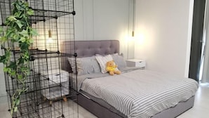A bedroom with a grey upholstered bed, striped bedding, a teddy bear, a bedside table, a lamp, and a black metal plant stand.