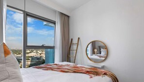 Habitación de hotel con un amplio ventanal que ofrece vista a la ciudad, una cama con colcha blanca, una escalera de madera y un espejo redondo en la pared.