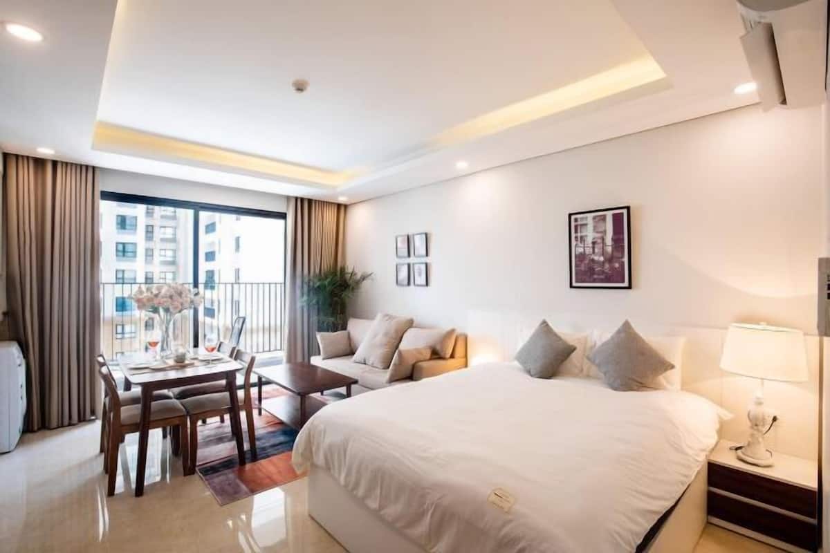 A modern hotel room with a large bed, a dining area, a sofa, and a balcony with curtains.