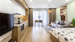 Una cocina moderna con armarios de madera y un televisor grande. Hay un comedor con dos sillas y una mesa. Se ve también un sofá y una cama con colcha estampada.