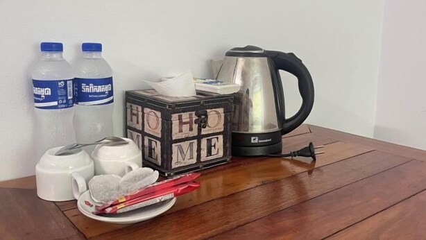 A wooden table with a black cube labeled "HO HO HO," two water bottles, a white teapot, a silver electric kettle, and a pair of scissors. - Le-nou Siemreap Guesthouse (Siem Reap)