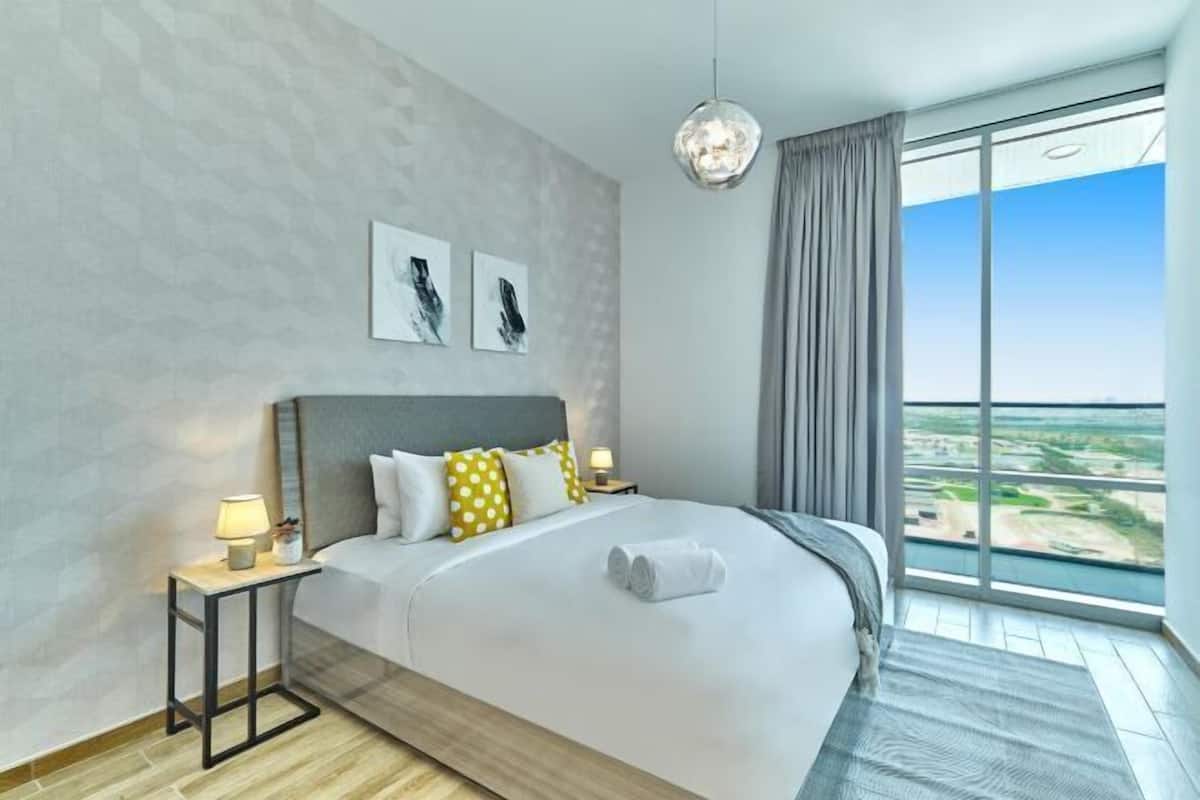A modern hotel room with a large bed, a nightstand, a lamp, a view of the city, and artwork on the wall.
