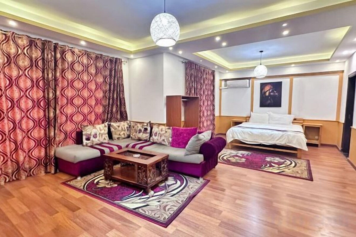 Une chambre d’hôtel avec un canapé d’angle, un lit, une table basse en bois et un tapis à motifs.