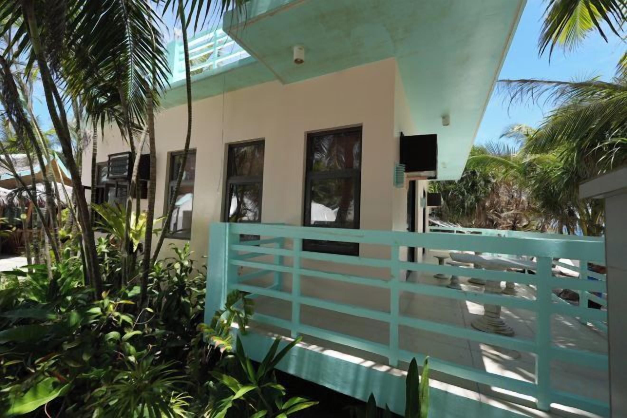 A building with a light-colored exterior, a blue fence, and tropical vegetation.