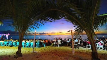 Un encuentro en la playa con gente sentada en sillas, palmeras y vista al atardecer.
