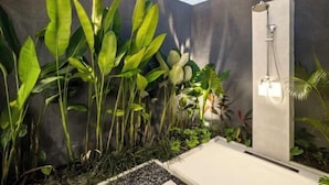 Une salle de bain moderne avec une douche, entourée d’une végétation luxuriante et d’un mur en béton.