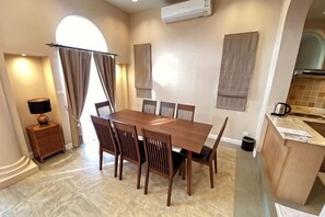 A dining area with a wooden table and chairs, a small side table with a lamp, and a view of the kitchen. - Villa Primo Khaoyai near Toscana (Pak Chong)