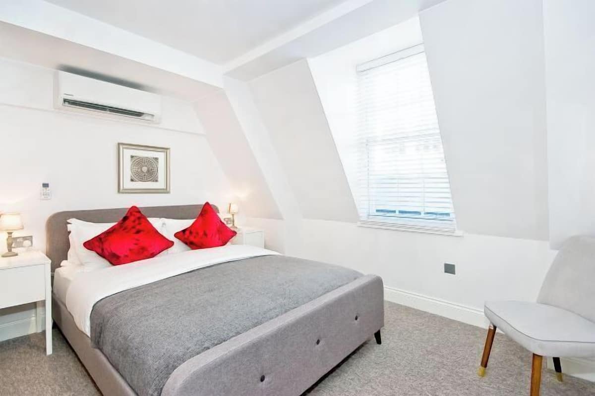 Un dormitorio con cama, mesita de noche, silla y ventana.