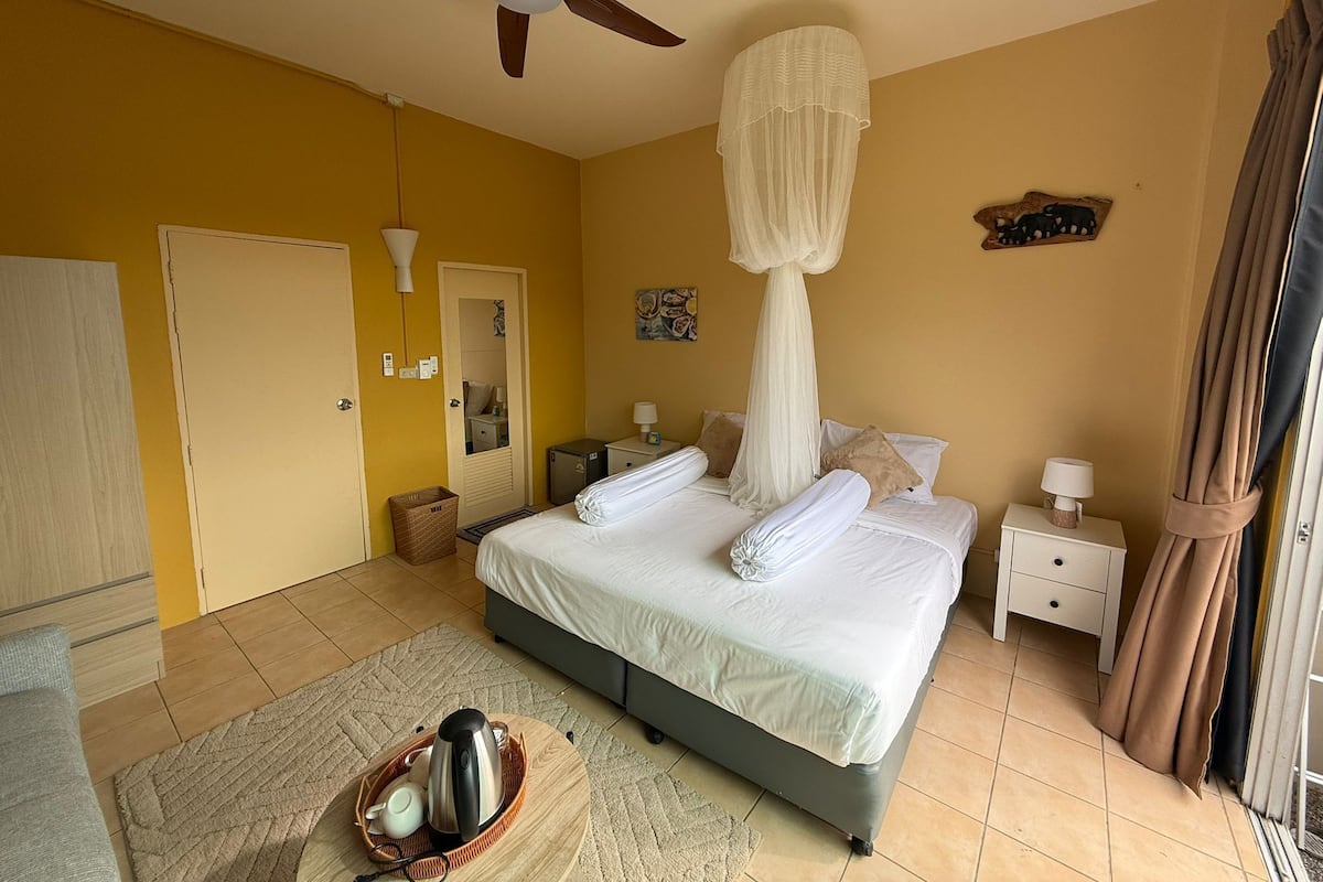 Une chambre à coucher comprenant un lit, des tables de chevet, un ventilateur de plafond, une fenêtre avec des rideaux et une porte.