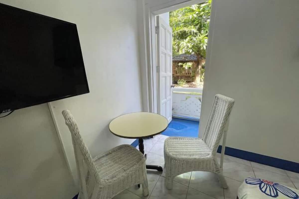 Une pièce équipée d’un téléviseur, de deux fauteuils en osier, d’une petite table ronde et offrant une vue sur un espace extérieur avec une piscine.