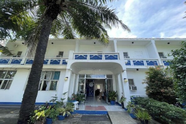 Un edificio de dos pisos con una entrada central, plantas en macetas y una palmera.