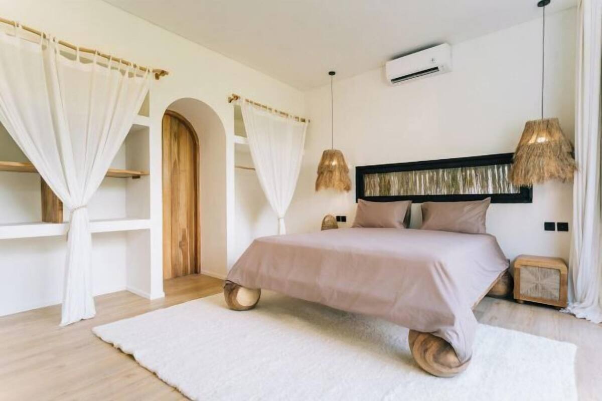 A bedroom with a bed, white curtains, a hanging light, and a small wooden side table.