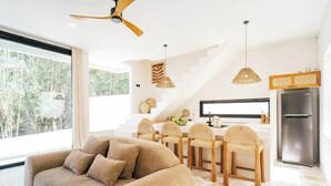 A modern living room with a sofa and dining area, large windows, and a kitchen with a refrigerator. - X Villa Khana · Exquisite 3br villa Canggu (Bali)
