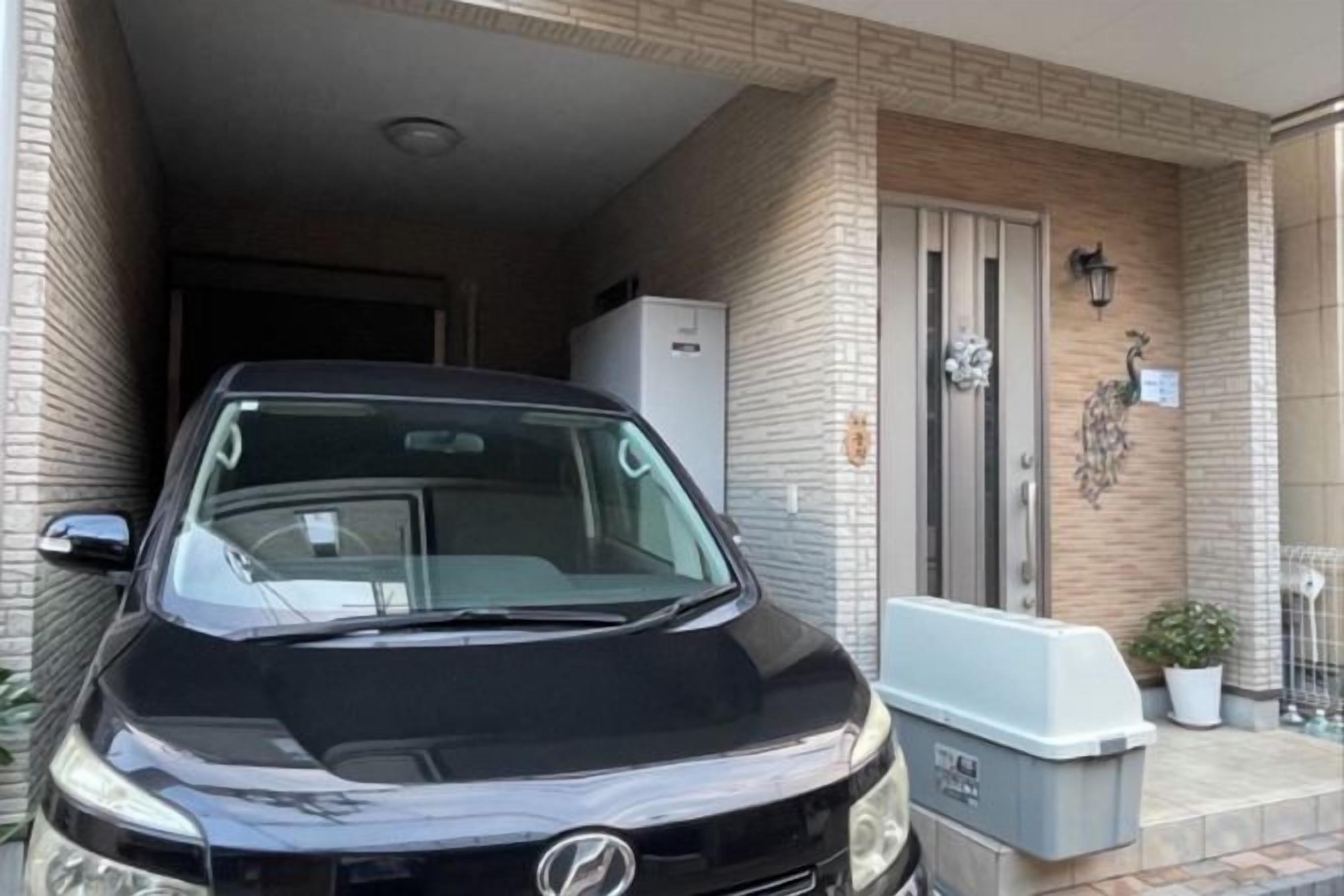 A black car parked in a covered parking area with a brick wall and a door to the left.