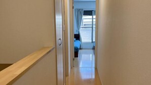 A narrow hallway with a wooden bench on the left, leading to a room with a bed and a window with curtains. - Peacock (Osaka)