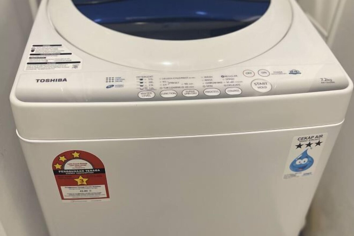 Une machine à laver blanche, dotée d’un panneau de commande frontal, d’un autocollant « CEKAP AIR » et d’une étiquette rouge ornée d’étoiles.