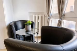 A modern living room with a black leather sofa, a glass table with a potted plant, and a view of a window with curtains. - huaxia (Venice)