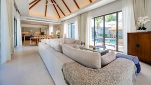 A modern living room with a large sofa, a coffee table, and a view of the outdoor pool area. - Anchan Tropicana Pool Villa near Laguna & Beach Phuket Warm Plus (Phuket)