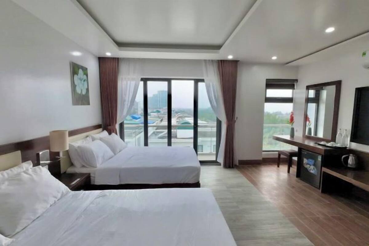 Ein Hotelzimmer mit zwei Betten, einem Fernseher, einem Schreibtisch und einem großen Fenster mit Blick auf Gebäude und ein Gewässer.
