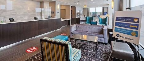 A modern hotel lobby with a reception desk, seating area, and a sign detailing COVID-19 safety measures.