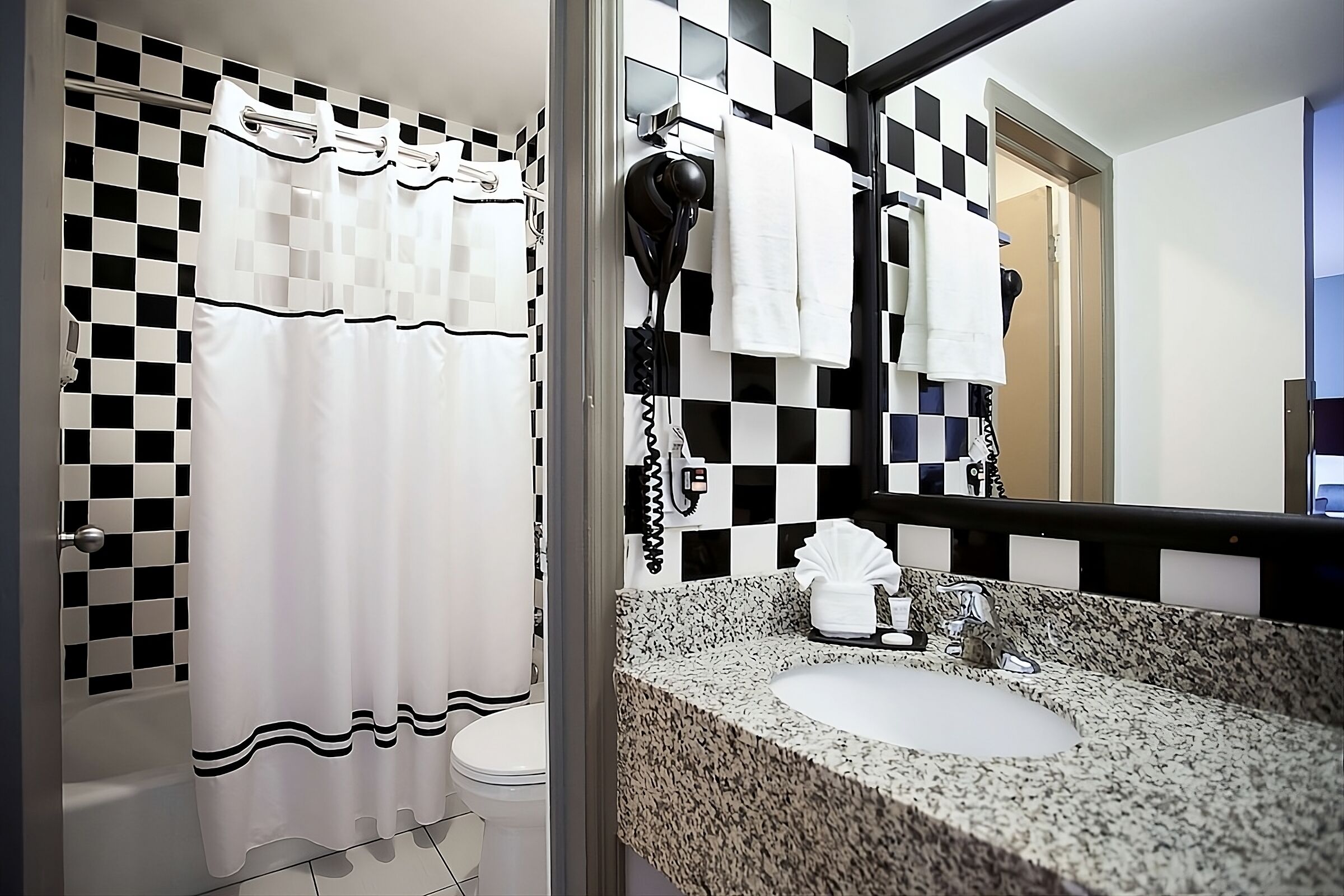 Une salle de bain avec un rideau de douche à carreaux, un sèche-cheveux fixé au mur et un lavabo sur un plan de travail en granit.