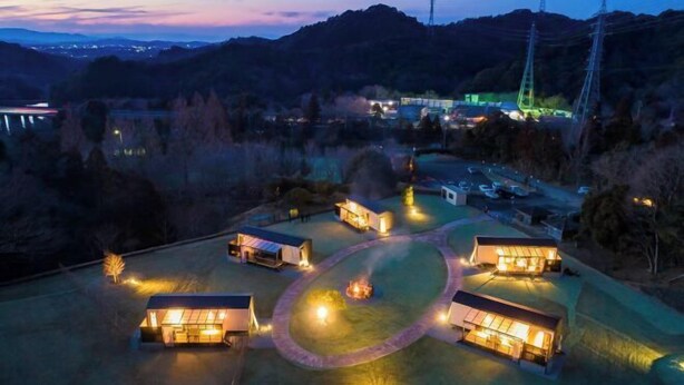A series of modern, well-lit cabins arranged in a circular pattern around a central fire pit, set in a mountainous area with visible power lines and distant buildings. - Luxury Trailers Rikyuise (Ise)