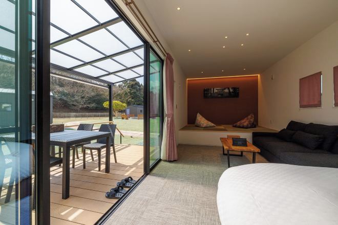 A modern room with a large bed, a seating area with a table and chairs, a glass wall with a view of the outdoors, and a cozy seating nook with cushions.