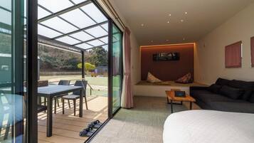 A modern room with a large bed, a seating area with a table and chairs, a glass wall with a view of the outdoors, and a cozy seating nook with cushions.