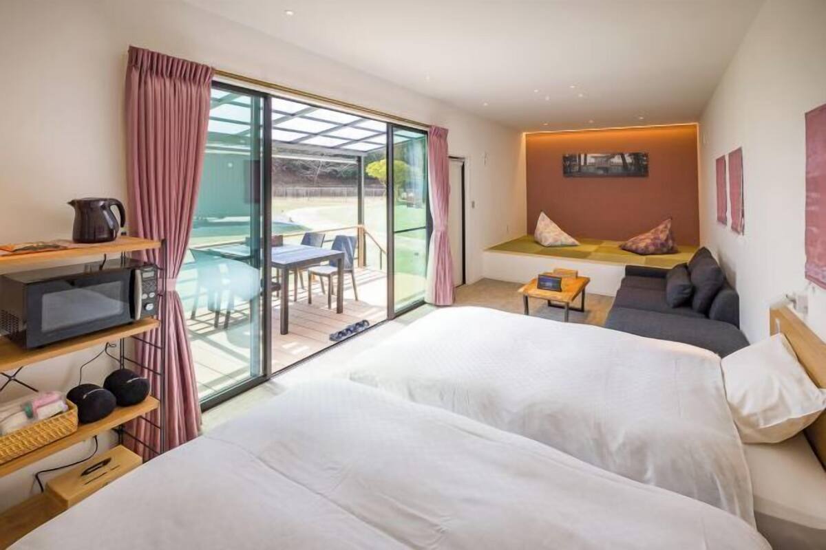 Quarto de hotel com duas camas, televisão, mesa pequena, sofá e porta de vidro deslizante para o exterior.