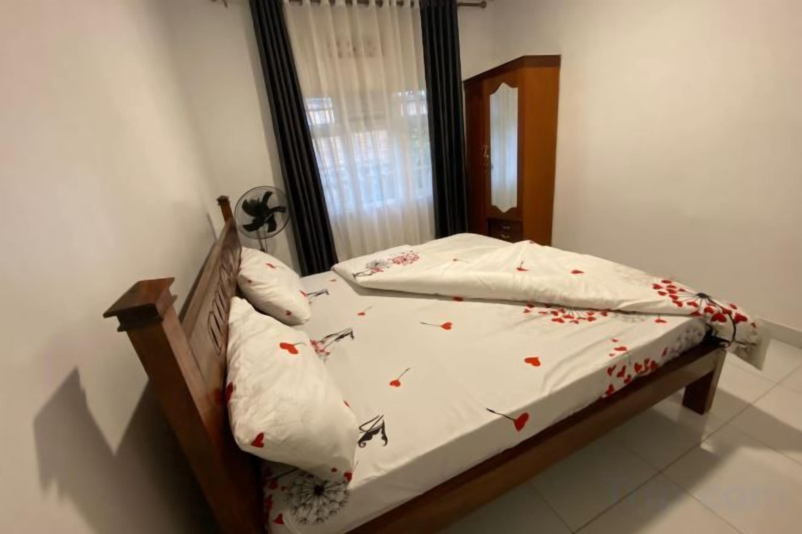 A single bed with a wooden frame, white bedding with red floral patterns, a bedside fan, and a window with curtains.