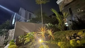 A nighttime view of a building with a modern design, featuring a stone wall and a well-lit garden with various plants. - Villa Sunrise F1 (Dau)
