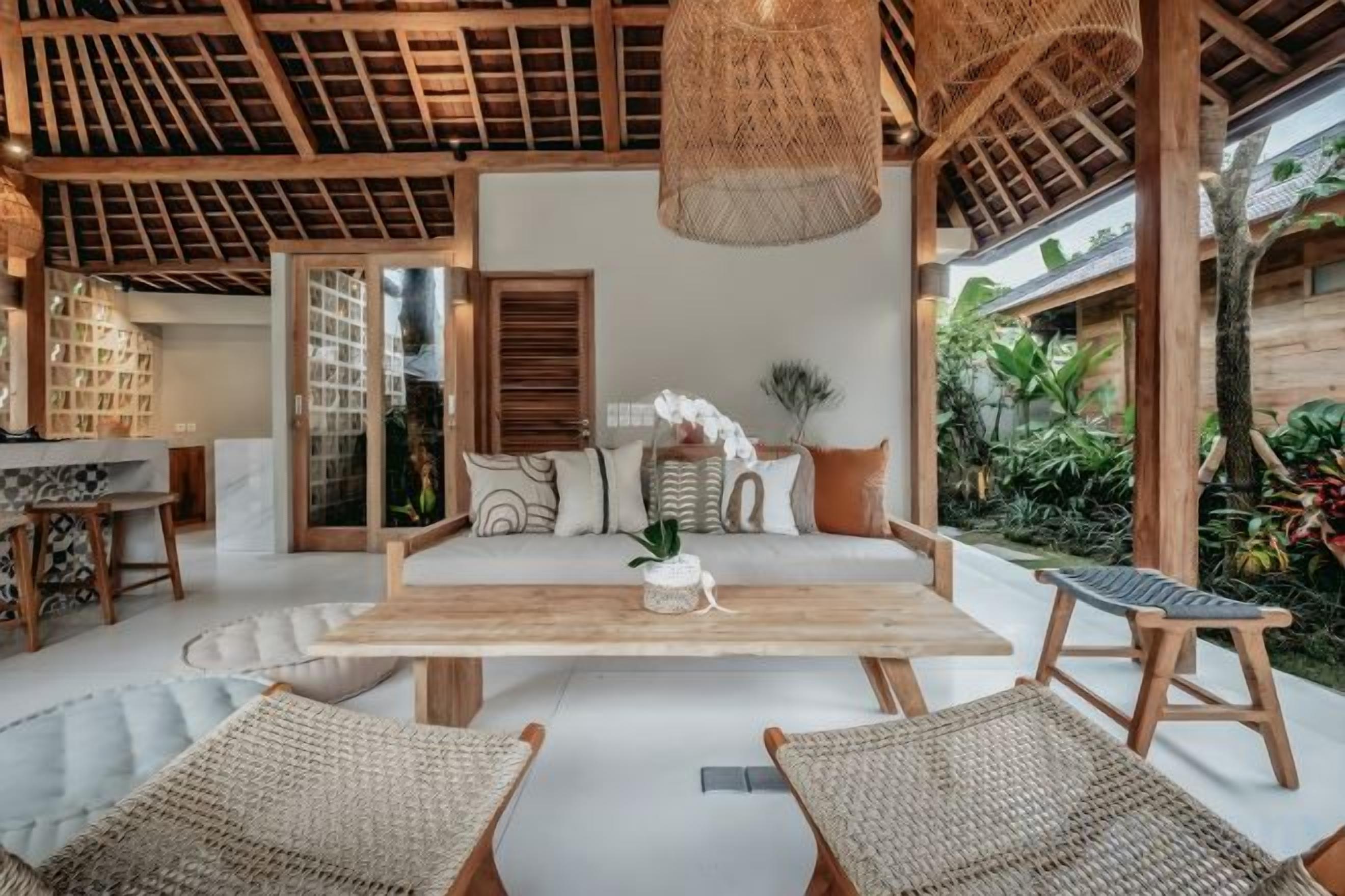 A room with a wooden ceiling, a central sofa, two wicker chairs, a hanging light fixture, and a view of greenery outside.