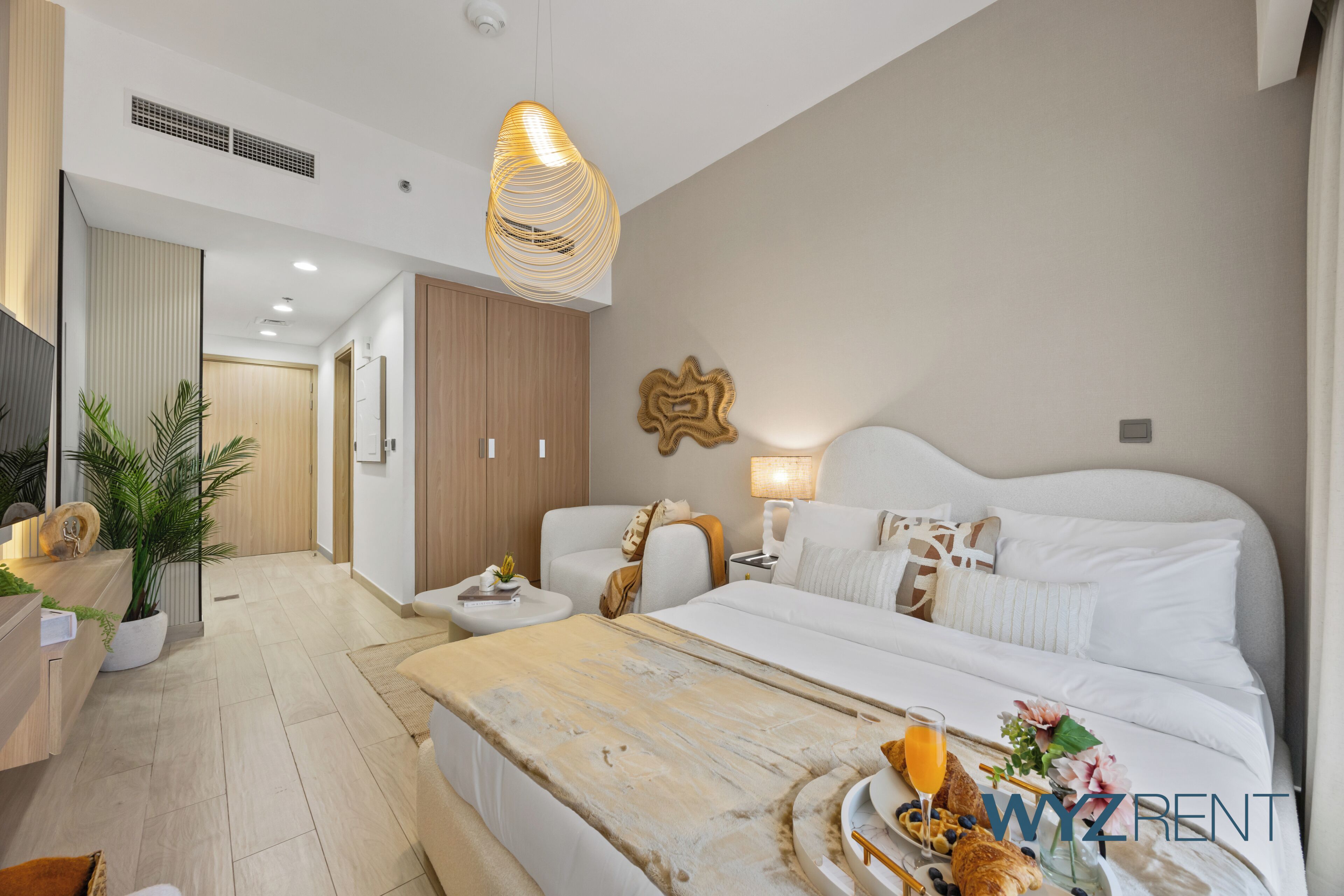 A modern hotel room with a large bed, a wooden coffee table, a desk with a plant, and a hanging light fixture.