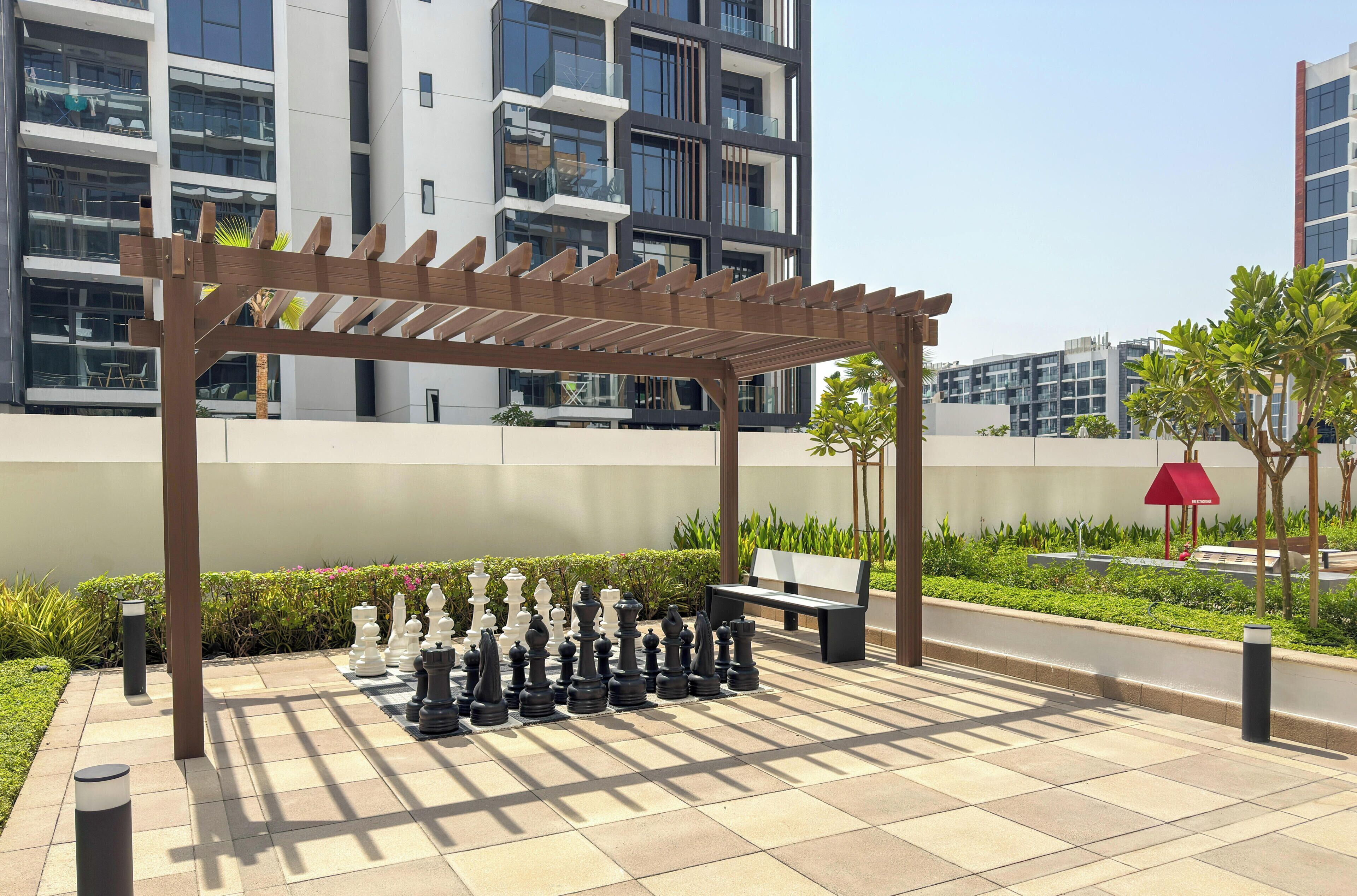 A chessboard set up on a tiled patio with a pergola overhead, surrounded by greenery and modern buildings in the background.