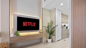 Un salon moderne doté d’un grand téléviseur à écran plat affichant le logo de Netflix, d’une console en bois agrémentée d’objets décoratifs, d’une plante en pot et d’une vue sur la cuisine adjacente.