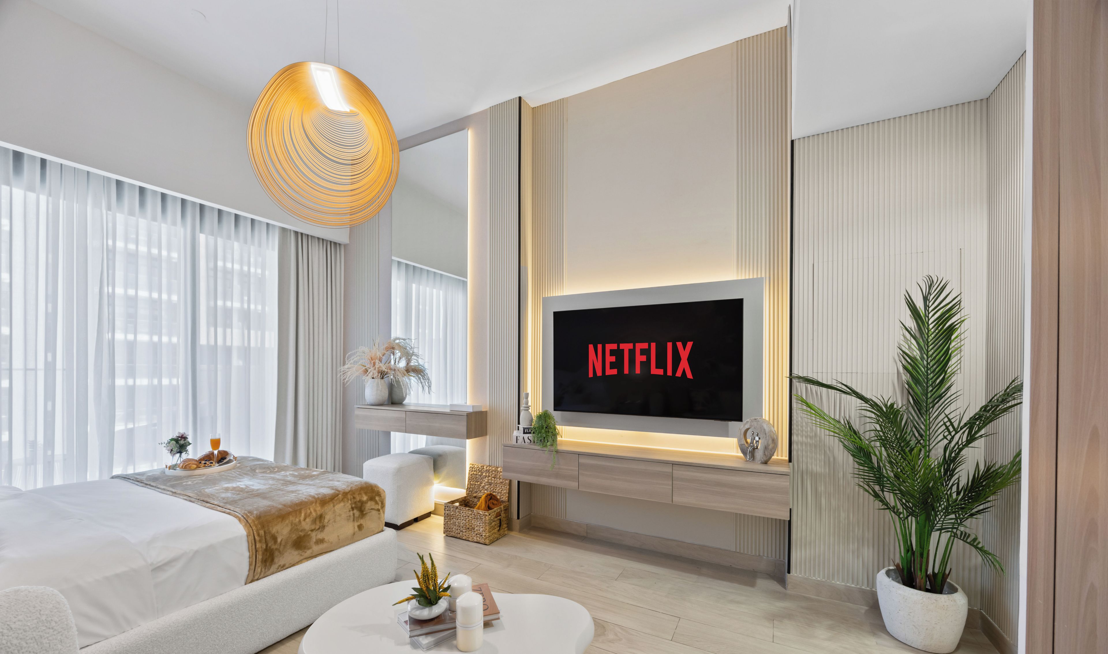 Una habitación de hotel moderna con una cama grande, un sofá blanco, una mesa de centro curvada, un televisor montado mostrando 'NETFLIX' y una planta en maceta.