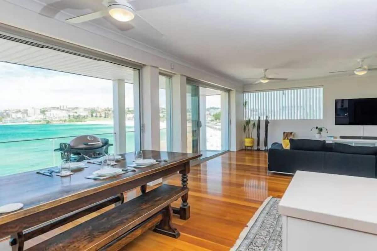 Amplia sala de estar con una mesa de comedor de madera, un sofá y un gran ventanal con vista al océano.