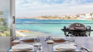 Une table à manger dressée pour quatre personnes, avec vue sur la mer et un barbecue en arrière-plan.