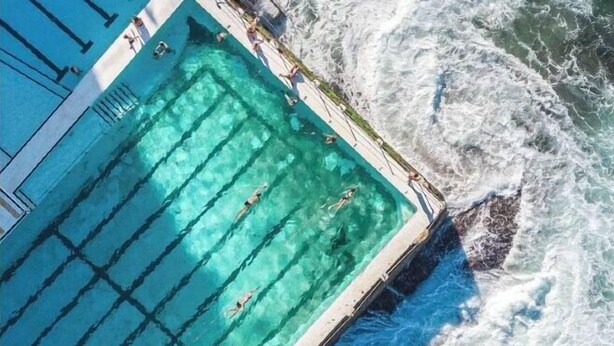 Une vue aérienne d’une piscine située à côté d’une côte rocheuse, où les vagues se brisent sur le rivage.
