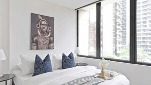A modern hotel room with a large bed, a window with a city view, a painting on the wall, and a nightstand with a lamp. - A Spacious 2BR Apt, Walk to Southern Cross Station (Melbourne)
