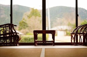 A room with a view of mountains through large windows, a tatami mat floor, and two wooden chairs.
