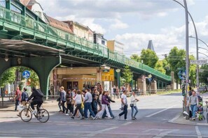 Een drukke stadsstraat met voetgangers, een fietser en een groene voetgangersbrug.
