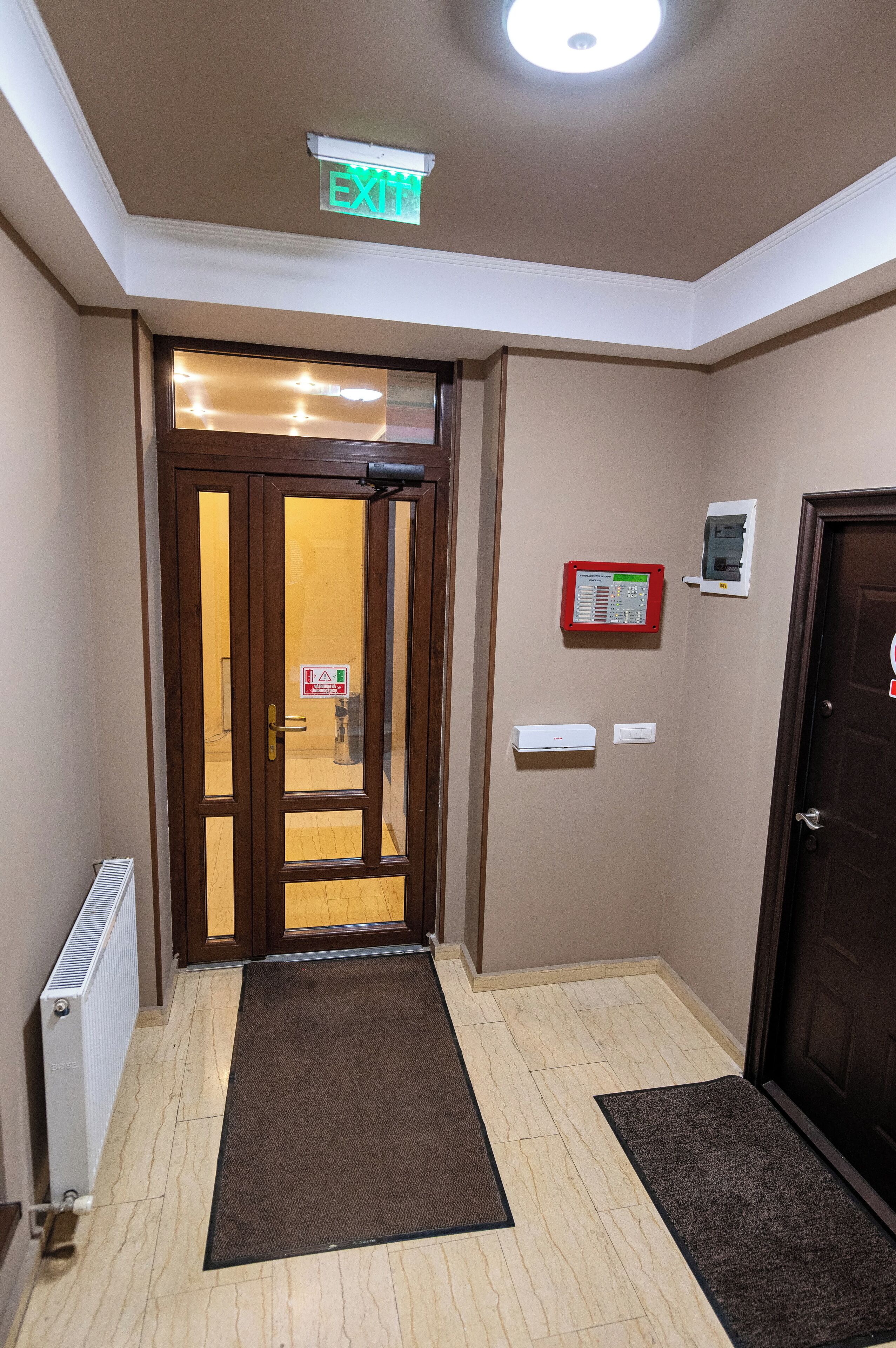 A hotel entrance with a brown door, a glass door, a sign indicating an exit, a fire alarm, and a radiator.