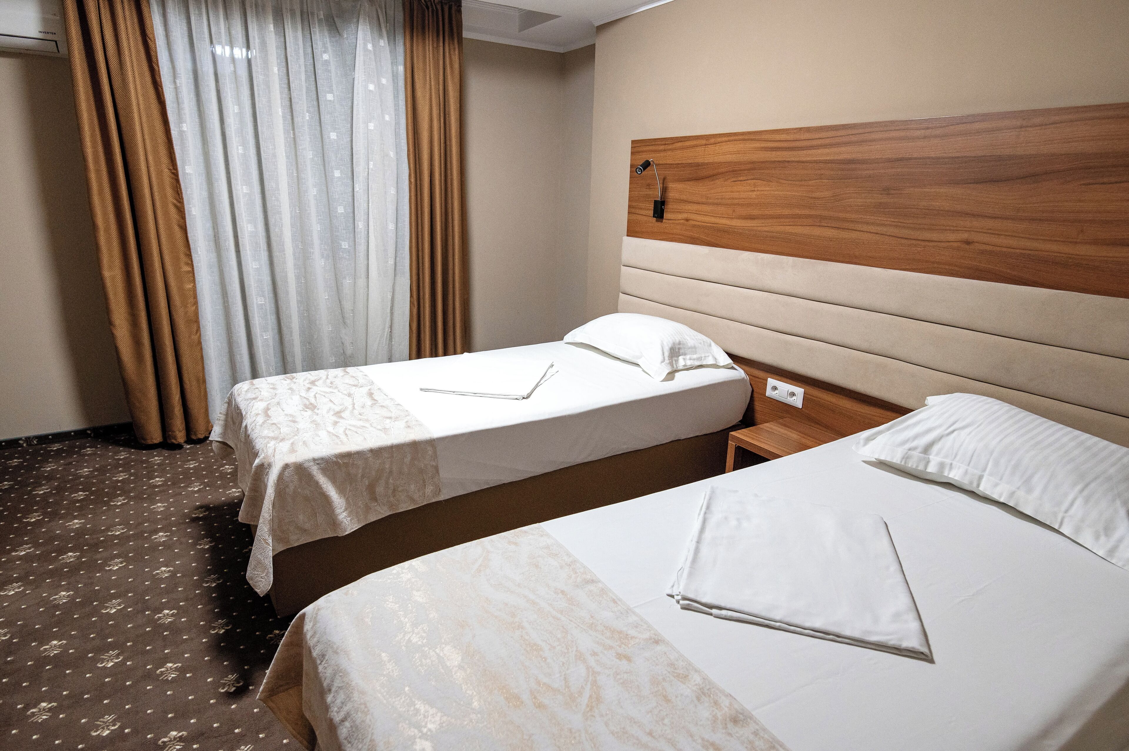 A hotel room with two beds, a wooden headboard, a bedside table, and a window with curtains.