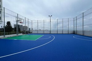 Una cancha de baloncesto interior con piso azul, rodeada por una valla de metal y un aro de baloncesto.