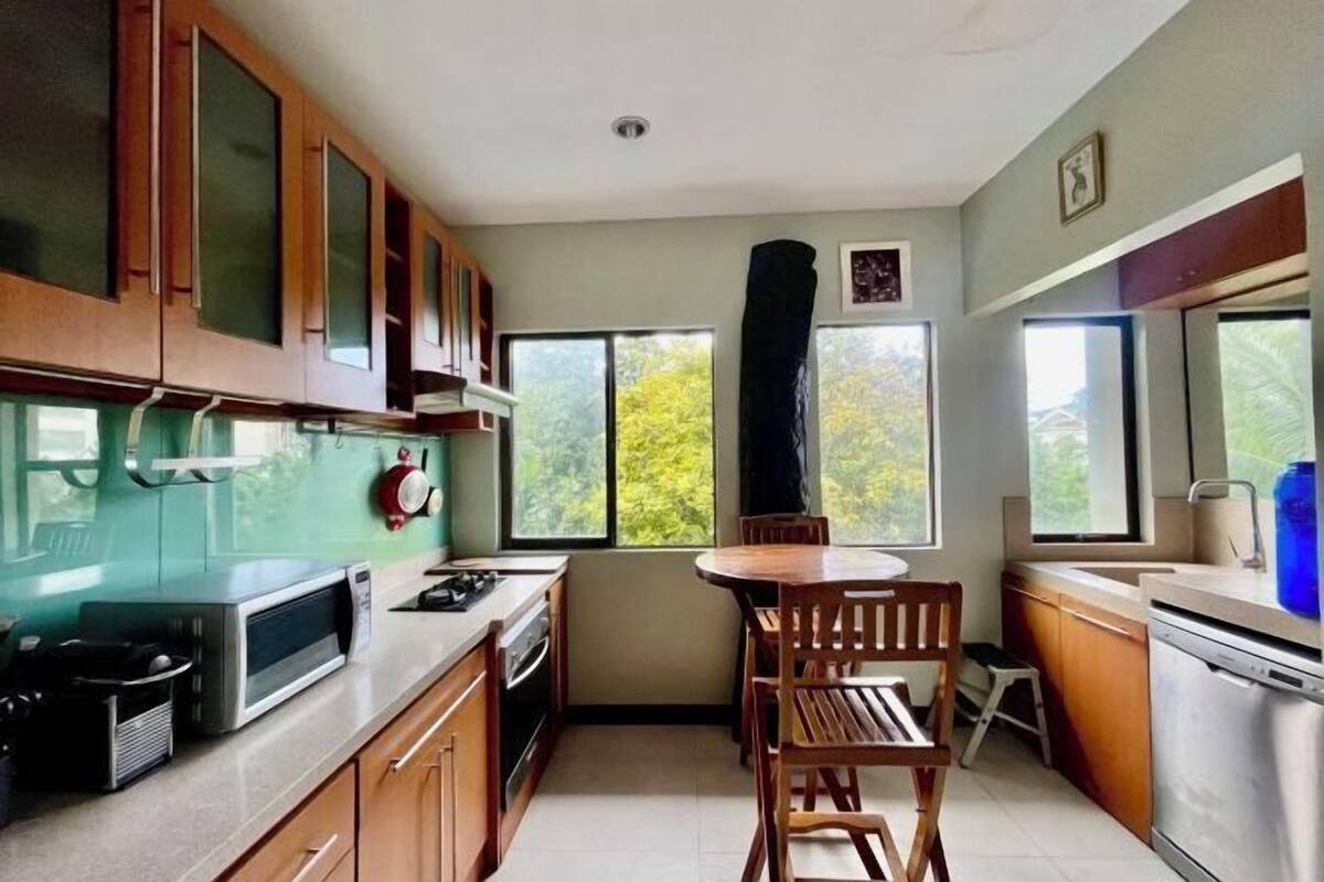 A compact kitchen with wooden cabinets, a microwave, a stove, and a sink. There is a window with a view of trees and a chair in the center.