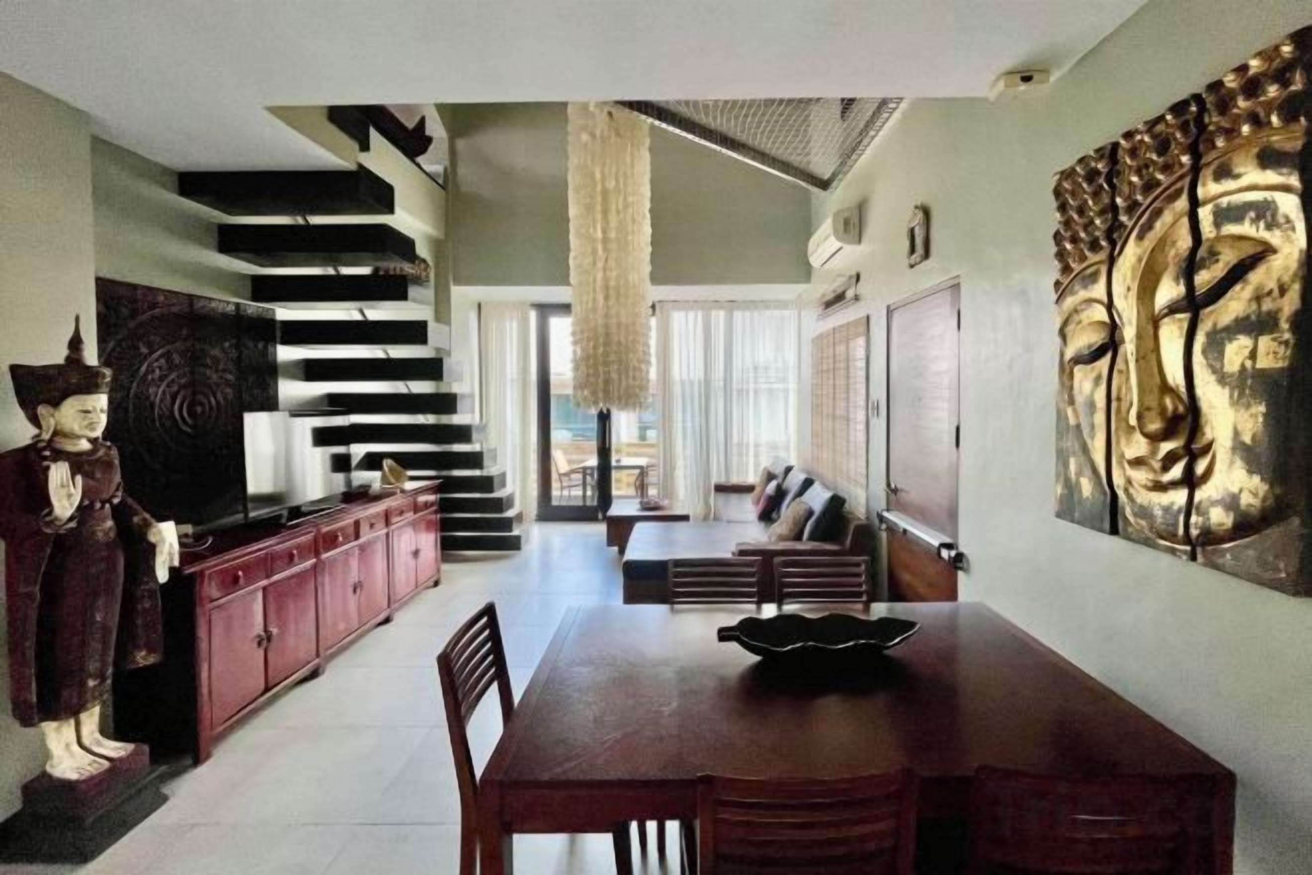 Un espace repas avec une longue table, des chaises et une statue. On y trouve un escalier avec une rampe et une statue de Bouddha accrochée au mur.