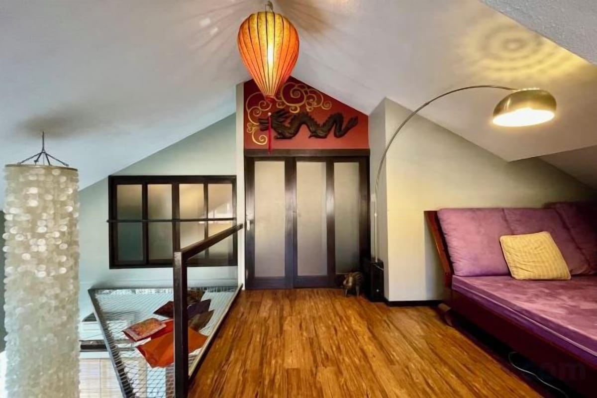A loft-style room with a sloped ceiling, a wooden floor, a purple sofa, a hanging light fixture, and a staircase leading to an upper level.
