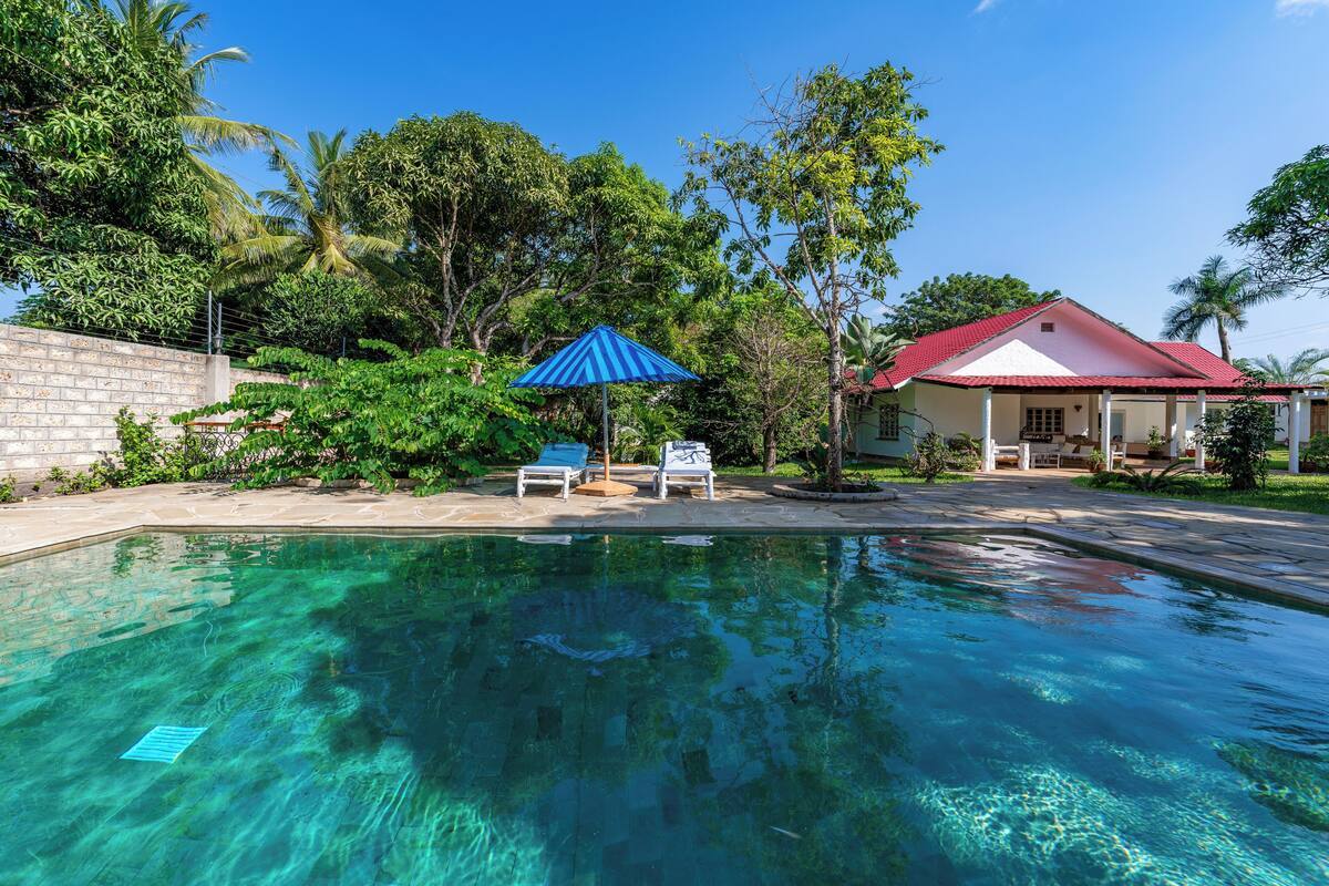 A clear swimming pool with two lounge chairs and a blue umbrella, surrounded by lush greenery and a building with a red roof.