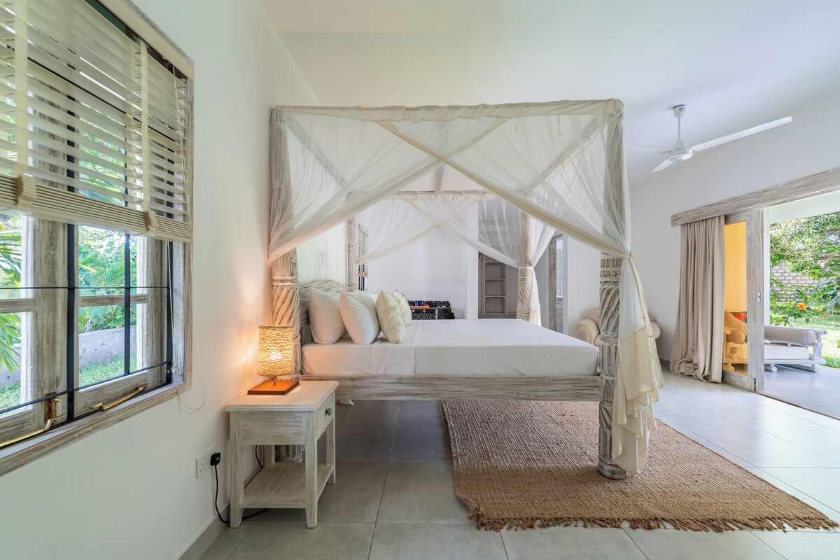A bedroom with a canopy bed, a bedside table with a lamp, a view of the outdoors, and a large window with blinds.